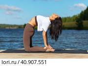 A woman practicing yoga, performs the Ushtrasana exercise, camel pose, trains in sportswear in leggings and a white t-shirt on a wooden bridge on the lake, sunny in the morning. Стоковое фото, фотограф Сергей Чайко / Фотобанк Лори