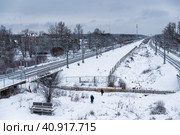Вид сверху на заснеженную железную дорогу. Электропоезд едет по железной дороге. Люди идут по снегу. Железнодорожная станция Саблино, деревня Ульяновка, Ленинградская область, Россия. Редакционное фото, фотограф Андрей Степанов / Фотобанк Лори