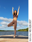 Woman practicing yoga performs Vrikshasana exercise, tree pose with arms raised up, balancing asana, exercising in sportswear on the shore of the lake on a warm sunny morning. Стоковое фото, фотограф Сергей Чайко / Фотобанк Лори