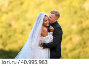 Счастливые молодожены обнимаются на фоне вечерней солнечной листвы, парень пытается весело укусить девушку. Стоковое фото, фотограф Иванов Алексей / Фотобанк Лори