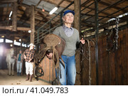 Elderly female rancher walking in stable with saddle in hands. Стоковое фото, фотограф Яков Филимонов / Фотобанк Лори