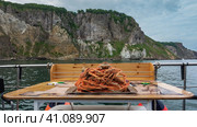 On the yacht, on the table, there is a tray. Boiled snow crabs are stacked in a slide. Legs and claws are visible. The background is the ocean, the rocky coast of Kamchatka. Стоковое фото, фотограф Вера Ивановна Кубарева / Фотобанк Лори