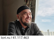 Portrait of happy senior asian man with beard wearing skullcap sitting by window and smiling. Стоковое фото, фотограф Ирина Кононова / Фотобанк Лори
