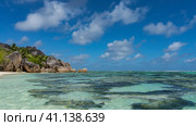 Corals on the bottom are visible through the clear turquoise water of the ocean. Fancy coastal cliffs with smoothed steep slopes against a blue sky, clouds. Sandy beach, tropical vegetation.Seychelles (2022 год). Стоковое фото, фотограф Вера Ивановна Кубарева / Фотобанк Лори