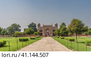 The entrance to the ancient tomb of Itmad-Ud-Daulah. The red sandstone walls are decorated with white ornaments. People walk along the path through the arched gate. Grass and trees on the roadsides (2022 год). Стоковое фото, фотограф Вера Ивановна Кубарева / Фотобанк Лори