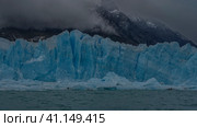 A wall of blue cracked ice rises above a turquoise lake. Ripples on the water. Snow-capped mountains are hiding in the fog. Argentina. Lago Argentino. Perito Moreno glacier. El Calafate. Стоковое фото, фотограф Вера Ивановна Кубарева / Фотобанк Лори