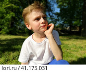 A boy looks into the distance and thinks. The child propped his chin in his hand and frowned. The process of thinking and a half smile. In the background is green grass and trees. Стоковое фото, фотограф Юлия Петровская / Фотобанк Лори
