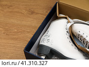 A pair of new white figure skates for women in the opened box. The box lies on a light brown wooden background. Selective focus. Copy space. Пара новых белых женских фигурных коньков в открытой коробке. Коробка лежит на светло-коричневом деревянном фоне. Избирательный фокус. Пространство для копирования. Стоковое фото, фотограф Дмитрий Березнев / Фотобанк Лори