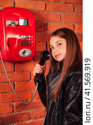 Little kid girl in leather jacket holding handset old red payphone at brick wall, looking at camera. Cute child lady model posing near telephone. Support communication concept. Copy ad text space. Стоковое фото, фотограф Aleksandr Golubev / Фотобанк Лори