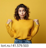 A young, pretty, attractive and thoughtful European girl of 20 years old in a gray casual T-shirt, arms spread apart, isolated on a uniform yellow background. Created by AI. Стоковое AI-изображение, AI-художник Ярославский Максим / Фотобанк Лори