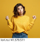 A young, pensive, insecure, rather attractive European girl of 20 years in a yellow casual blouse, spreads her hands questioningly, looking away. Created by AI. Стоковое AI-изображение, AI-художник Ярославский Максим / Фотобанк Лори