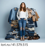 A young attractive girl of about 20 years old is standing in a blouse and jeans in front of a large pile of clothes on a white background. Generated by AI. Стоковое AI-изображение, AI-художник Ярославский Максим / Фотобанк Лори