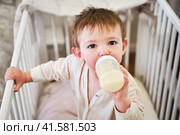 Toddler baby is standing with a bottle of formula milk in the crib, portrait happy child. Kid is a boy aged one year and three months. Стоковое фото, фотограф Андрей Журавлев / Фотобанк Лори