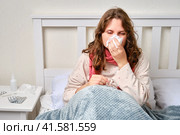 A sick woman blows her nose in a napkin, a runny nose. Adult ill woman with a red scarf sitting on a home bed in a white bedroom, female aged 35 years. Стоковое фото, фотограф Андрей Журавлев / Фотобанк Лори