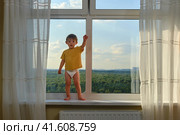 Baby boy is standing on the high window sill, risking their safety. Kid aged two years. Стоковое фото, фотограф Андрей Журавлев / Фотобанк Лори