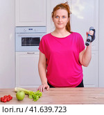 Young woman in home kitchen with dumbbells, vegetables and fruit. Concept of proper nutrition and sports in isolation during the coronavirus epidemic. Стоковое фото, фотограф Андрей Журавлев / Фотобанк Лори