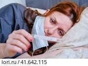 A woman with a coronavirus looks sadly at a thermometer. The girl lies on the bed in a medical mask. Стоковое фото, фотограф Андрей Журавлев / Фотобанк Лори