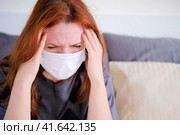 A woman with coronavirus in a medical mask holds her head, copy space for text. A girl with a sore head is sitting on the bed. Стоковое фото, фотограф Андрей Журавлев / Фотобанк Лори
