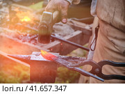 The blacksmith has a hammer on hot iron. The workpiece is clamped in the grip of the master. Bellows with fire on the background of handicraft work. Стоковое фото, фотограф Андрей Журавлев / Фотобанк Лори