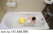 Baby drinks water from a ladle while sitting with toys in a white bathtub A happy child enjoys washing in the bathroom. Kid about two years old (one year ten months). Стоковое видео, видеограф Андрей Журавлев / Фотобанк Лори