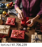 An unrecognizable woman wraps Christmas gifts in red paper. Стоковое AI-изображение, AI-художник Margarita Davchenko / Фотобанк Лори