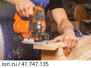 Close up of experienced carpenter in work clothes and small buiness owner  carpenter saw and processes the edges of a wooden bar with a jig saw  in a workshop. Стоковое фото, фотограф Виталий Сова / Фотобанк Лори