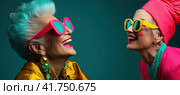 Two elderly beautiful women with brightly colored hair, wearing sunglasses smile and look up on a blue background. Generated by AI. Стоковое AI-изображение, AI-художник Ярославский Максим / Фотобанк Лори