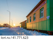 Село Тавайваам, Чукотка, Россия. Вид на заснеженную улицу арктического посёлка и здание детского сада. На стене вывеска «Детский сад Оленёнок» и табличка с названием улицы «Колхозная». Редакционное фото, фотограф Андрей Степанов / Фотобанк Лори