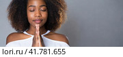 Young African American woman praying and hoping for good luck. Girl in white t-shirt begs God to hear her prayer. Female portrait with folded hands. Gray background with copy space. Panorama. Стоковое AI-изображение, AI-художник Igor Tichonow / Фотобанк Лори
