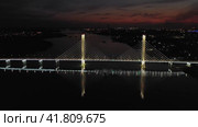 A cable-stayed highway bridge with tall concrete pylons illuminated by architectural lighting at night. A panorama of the riverbed running away under the night sky against the background of city lights (2023 год). Редакционное видео, видеограф Даниил Хабаров / Фотобанк Лори