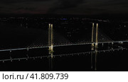 A cable-stayed highway bridge with tall concrete pylons illuminated by architectural lighting at night. General plan of the bridge against the background of night city lights, flying backward (2023 год). Редакционное видео, видеограф Даниил Хабаров / Фотобанк Лори