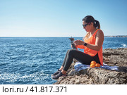 Latin woman, middle-aged, resting, regaining strength, eating, drinking water, after a gym session, burning calories, keeping fit, outdoors by the sea, wearing headphones and smart watch. Стоковое фото, фотограф carlos l vives / Фотобанк Лори