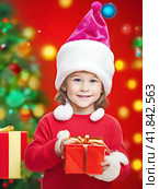 Smiling boy in a red Christmas hat with gifts near the Christmas tree. Стоковое AI-изображение, AI-художник Сергей Елизарьев / Фотобанк Лори
