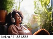 Chubby young dark skinned women sits in relaxed posture listening music with headphones among plants. Стоковое AI-изображение, AI-художник Наталья Семкина / Фотобанк Лори