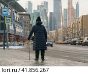 Aged woman with crutch walks on the Moscow city street. Old houses are combined with modern buildings. Walk with a limp in the city center. Back view. Стоковое фото, фотограф YuryKara / Фотобанк Лори