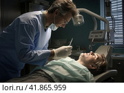 A frightened man lies in a dental chair, the dentist leans over him to treat his teeth. Pathological fear of dental treatment. Стоковое AI-изображение, AI-художник Коханова Наталья / Фотобанк Лори
