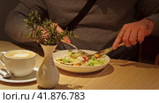 Young Man Wearing Gray Sweater Eating Fresh Vegetable Salad. Unrecognized Male Person Eating Caesar Salad in a Cafe or Restaurant and drinking Coffee while Sitting at a Wooden Table. Business Lunch. Стоковое видео, видеограф Марина Демидюк / Фотобанк Лори
