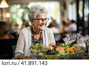 Portrait of a senior woman in a retirement home happily enjoying a healthy lunch. Presentation of a healthy lifestyle of well-being and contentment even at an age. Стоковое AI-изображение, AI-художник Ирина Михайличенко / Фотобанк Лори