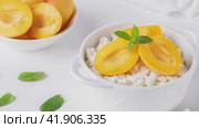 Fresh cottage cheese in white bowl with mint leaves and peach pieces and teaspoon on white background. High quality 4k footage. Стоковое видео, видеограф Наталия Попова / Фотобанк Лори