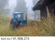 An old blue agricultural tractor in the tall grass on the outskirts of the village. Morning fog. Summer rural landscape. Стоковое фото, фотограф Андрей Степанов / Фотобанк Лори