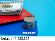 Карты платежных систем Мир и Виза с пачкой долларов и стопкой рублевых монет на голубом фоне. Место для надписи. (2024 год). Редакционное фото, фотограф Ярославский Максим / Фотобанк Лори