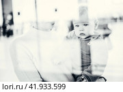 Thoughtful young mother looking trough window holding his infant baby boy child while waiting to board an airplane at airport terminal departure gates. Travel with baby concept. Стоковое фото, фотограф Matej Kastelic / Фотобанк Лори