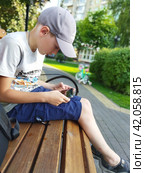 A cute boy on a board bench in a well-equipped city park. The kid about nine in light summer clothes looks attentively at a smartphone. A small planted square looks good in warm late afternoon light (2023 год). Редакционное фото, фотограф Анатолий Власов / Фотобанк Лори