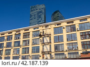 Вид на стену промышленного здания, за которой возвышаются небоскрёбы. Здания на фоне синего неба. Редакционное фото, фотограф Андрей Степанов / Фотобанк Лори