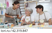 Friendly older mother and young adult daughter and son, wearing aprons, talking cheerfully while working on painting ceramic dishware in family pottery workshop. Generational collaboration concept. Стоковое видео, видеограф Яков Филимонов / Фотобанк Лори