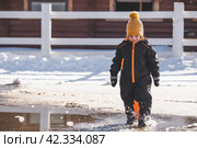 Portrait of a little boy in a yellow hat. The child runs through puddles on the farm, has fun and plays. It's a sunny day. Early spring. The first drop. Стоковое фото, фотограф Photojuli / Фотобанк Лори