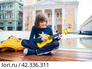 10 летний мальчик играет на желтой укулеле, пластиковой гавайской маленькой гитаре, сидя у фонтана на Биржевой площади в Москве. Стоковое фото, фотограф Яна Проничева / Фотобанк Лори
