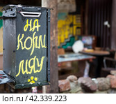 Ящик с надписью "На корм мяу" на фоне кушающей кошки. «Кошкин двор». Гвардейск, Калининградская область (2023 год). Редакционное фото, фотограф Сергей Афанасьев / Фотобанк Лори