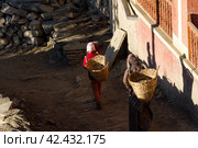 Jharkot, Nepal - November 18, 2015: Nepalese women carry on their back baskets for cargo along the street in the village in Nepal. Редакционное фото, фотограф VittoriaChe / Фотобанк Лори