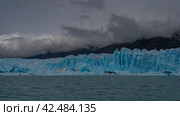 A wall of blue ice with cracks, crevices, and sharp peaks stretches in a glacial turquoise lake. Thawed ice floes, icebergs floating in the water. Mountains in clouds and fog. Perito Moreno glacier. Стоковое фото, фотограф Вера Ивановна Кубарева / Фотобанк Лори